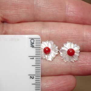 May include: A pair of flower-shaped earrings. Each earring features a white, petal-like design surrounding a small, round, red center. The earrings are being measured with a ruler, and are approximately 1 cm in diameter.