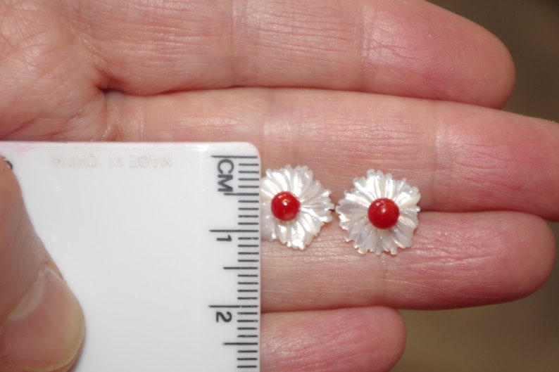 May include: A pair of flower-shaped earrings. Each earring features a white, petal-like design surrounding a small, round, red center. The earrings are being measured with a ruler, and are approximately 1 cm in diameter.
