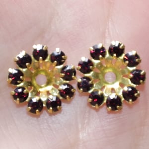 Puede incluir: Un par de cierres de pendientes de metal dorado con una base circular y un anillo en el centro. El anillo está rodeado de 12 pequeños diamantes de imitación de color rojo oscuro.