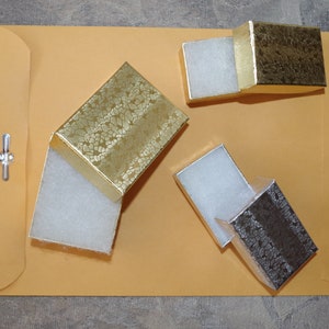 May include: Three small, open gift boxes with textured gold and silver lids, each containing white padding, are arranged on a large, tan envelope. The boxes are in various positions, showcasing their interior. The envelope has a metal clasp.
