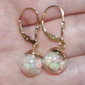 May include: A pair of gold-colored leverback earrings. Each earring features a clear, round glass orb filled with small, white and iridescent opal stones. The earrings are displayed against a soft, neutral background.