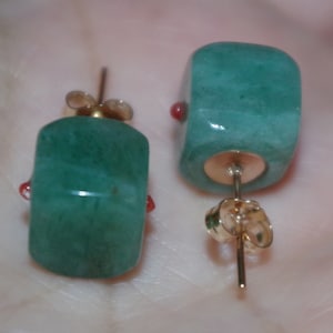 May include: A pair of green gemstone earrings with red accents and gold posts. The earrings are square shaped and have a smooth, polished finish.
