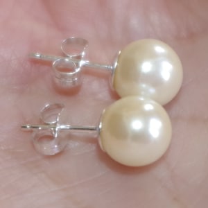 May include: A pair of cream-colored pearl stud earrings with silver posts and backs. The earrings are round and have a glossy finish, reflecting light. The posts are straight and have a butterfly clasp.