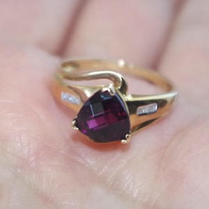 Könnte beinhalten: Ein goldener Ring mit einem herzförmigen, violetten Edelstein und kleinen Diamanten auf der Ringschiene.