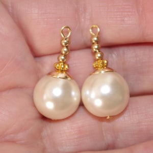 May include: Two cream-colored pearl earrings with gold-colored accents. Each earring features a large, round pearl, topped with a gold-colored cap, a string of small gold beads, and a gold loop for hanging.
