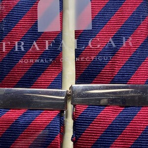 May include: Two striped neckties with a red and navy blue pattern, held together by a silver clasp. The brand name "TRAFALGAR" and the city "NORWALK, CONNECTICUT" are visible on the packaging.