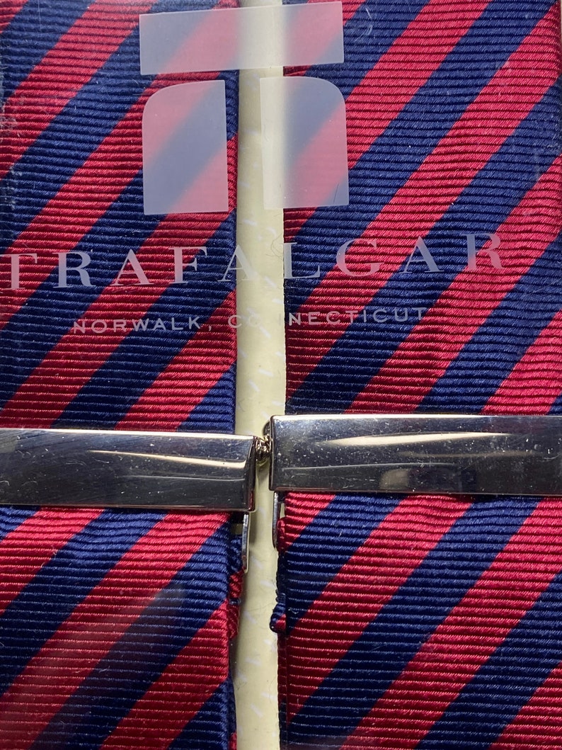 May include: Two striped neckties with a red and navy blue pattern, held together by a silver clasp. The brand name "TRAFALGAR" and the city "NORWALK, CONNECTICUT" are visible on the packaging.