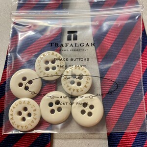 May include: A package of Trafalgar brace buttons with the text "BRACE BUTTONS" and "TRAFALGAR NORWALK, CONNECTICUT" printed on the packaging. The buttons are ivory-colored and have the brand name printed on them. A red and blue striped tie is in the background.