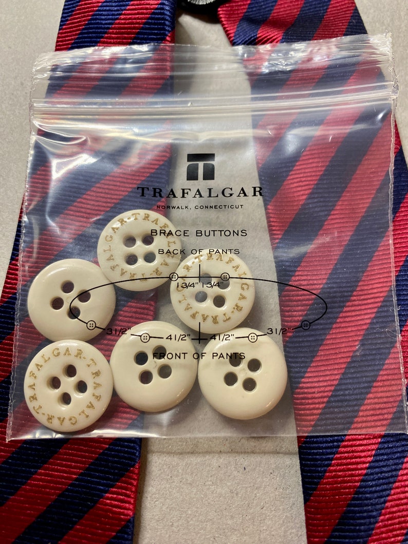 May include: A package of Trafalgar brace buttons with the text "BRACE BUTTONS" and "TRAFALGAR NORWALK, CONNECTICUT" printed on the packaging. The buttons are ivory-colored and have the brand name printed on them. A red and blue striped tie is in the background.