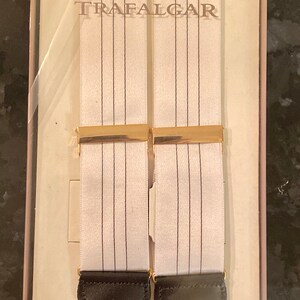 May include: White suspenders with thin black vertical stripes and gold-colored metal clasps. The suspenders have black leather ends and the word "TRAFALGAR" is printed on the packaging.