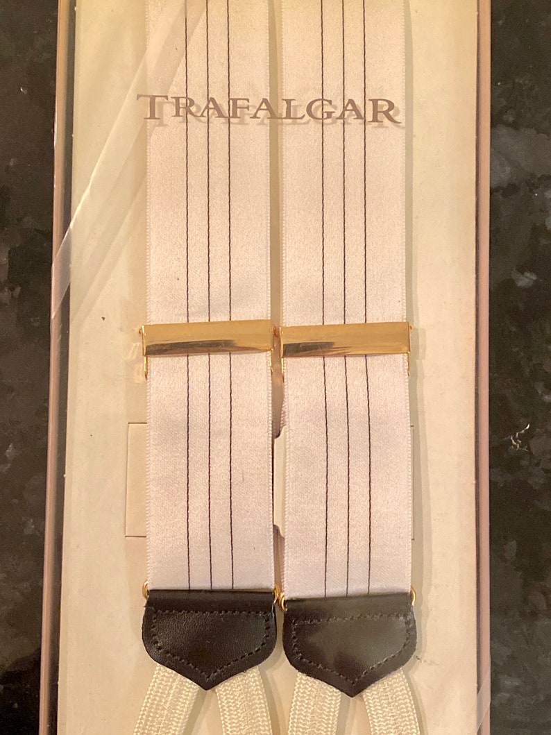 May include: White suspenders with thin black vertical stripes and gold-colored metal clasps. The suspenders have black leather ends and the word "TRAFALGAR" is printed on the packaging.