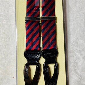 May include: Red and navy blue striped suspenders with black leather ends and silver-tone metal clasps. The suspenders are packaged in a clear plastic box with a tan backing. The brand name "Trafalgar" is visible on the suspenders.