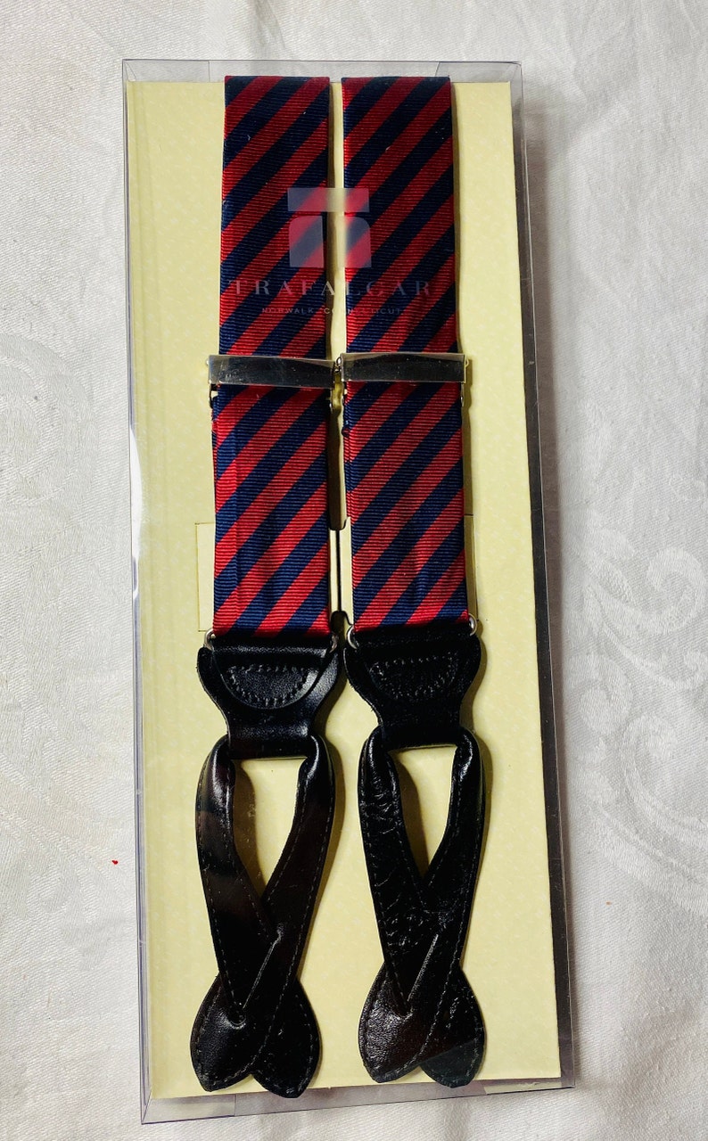 May include: Red and navy blue striped suspenders with black leather ends and silver-tone metal clasps. The suspenders are packaged in a clear plastic box with a tan backing. The brand name "Trafalgar" is visible on the suspenders.