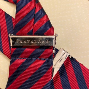 May include: Close-up of a pair of suspenders with a red and navy blue striped pattern. The metal clasp is engraved with the word "TRAFALGAR". The suspenders are made of a ribbed fabric.