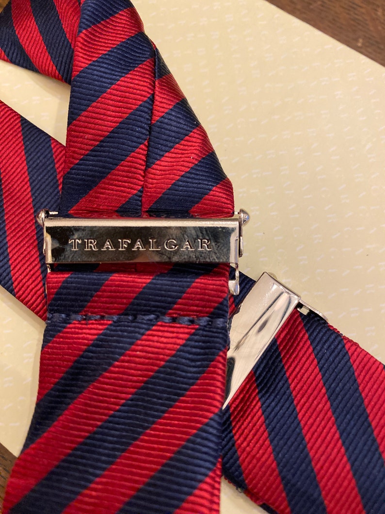 May include: Close-up of a pair of suspenders with a red and navy blue striped pattern. The metal clasp is engraved with the word "TRAFALGAR". The suspenders are made of a ribbed fabric.