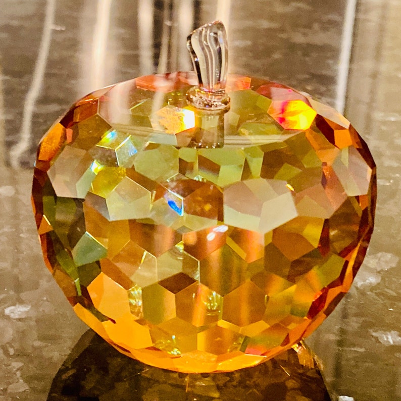 K&ouml;nnte beinhalten: Ein dekorativer Apfel aus facettiertem Glas in Orange-, Gelb- und Gr&uuml;nt&ouml;nen. Er hat einen klaren, gebogenen Stiel. Die Oberfl&auml;che besteht aus geometrischen Formen. Der Apfel steht auf einer reflektierenden Oberfl&auml;che.