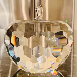 K&ouml;nnte beinhalten: Eine klare, facettierte Kristallapfel-Skulptur mit Stiel. Die Oberfl&auml;che des Apfels besteht aus zahlreichen geometrischen Formen, die das Licht reflektieren und einen Regenbogeneffekt erzeugen. Der Stiel ist ebenfalls aus klarem Kristall.