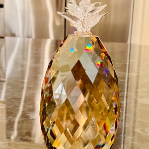 K&ouml;nnte beinhalten: Eine dekorative Kristall-Ananas-Figur. Der Ananask&ouml;rper hat eine facettierte bernsteinfarbene Farbe mit einer klaren, strukturierten Oberseite. Der Kristall reflektiert das Licht und erzeugt einen Regenbogeneffekt. Ein dekoratives Objekt.