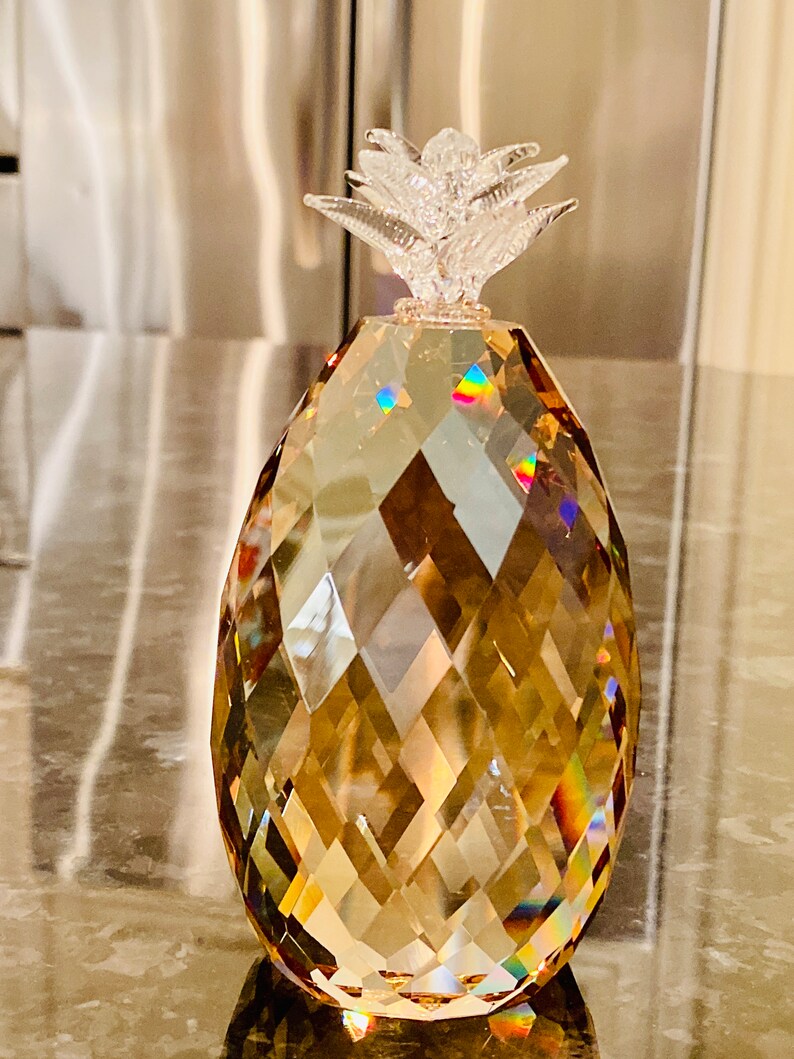 K&ouml;nnte beinhalten: Eine dekorative Kristall-Ananas-Figur. Der Ananask&ouml;rper hat eine facettierte bernsteinfarbene Farbe mit einer klaren, strukturierten Oberseite. Der Kristall reflektiert das Licht und erzeugt einen Regenbogeneffekt. Ein dekoratives Objekt.