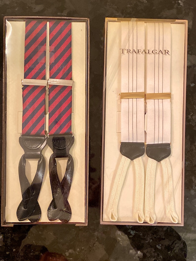 May include: Two pairs of suspenders in boxes. One pair has red and navy blue diagonal stripes. The other pair is white with thin black vertical stripes. Both pairs have leather ends and metal clasps.