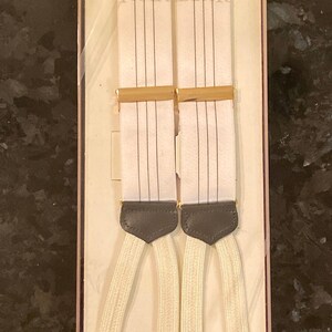 May include: White suspenders with thin black vertical stripes, packaged in a box. The suspenders have cream-colored elastic straps and dark gray leather accents. The word "TRAFALGAR" is printed on the box.