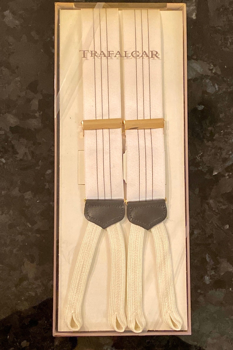 May include: White suspenders with thin black vertical stripes, packaged in a box. The suspenders have cream-colored elastic straps and dark gray leather accents. The word "TRAFALGAR" is printed on the box.