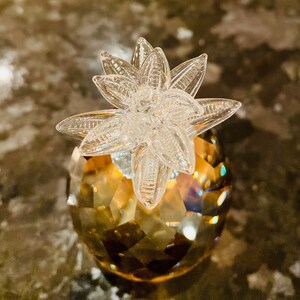 K&ouml;nnte beinhalten: Dekorative Kristall-Ananas-Figur. Der Ananask&ouml;rper ist facettiert und hat einen goldbraunen Farbton, w&auml;hrend die Oberseite ein klares, sternf&ouml;rmiges Design aufweist. Die Figur ist ein dekoratives Objekt.