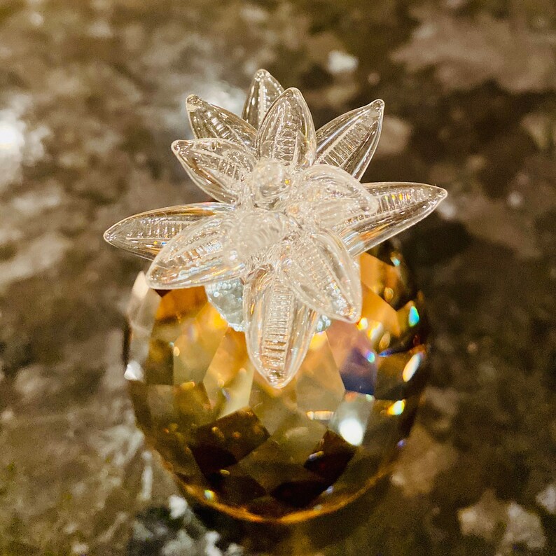 K&ouml;nnte beinhalten: Dekorative Kristall-Ananas-Figur. Der Ananask&ouml;rper ist facettiert und hat einen goldbraunen Farbton, w&auml;hrend die Oberseite ein klares, sternf&ouml;rmiges Design aufweist. Die Figur ist ein dekoratives Objekt.