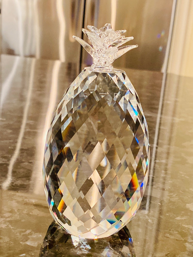 K&ouml;nnte beinhalten: Eine klare, facettierte Kristall-Ananas-Figur. Die Ananas hat einen strukturierten K&ouml;rper und eine detaillierte, blattartige Krone. Der Kristall reflektiert das Licht und erzeugt einen Regenbogeneffekt. Dieser Dekorationsartikel ist ein einzigartiges Wohnaccessoire.