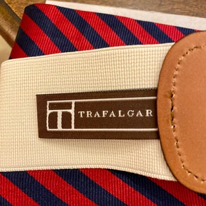 May include: Close-up of a striped belt with the word "TRAFALGAR" printed on a brown label. The belt features red and navy blue diagonal stripes, a beige elastic band, and a brown leather detail.