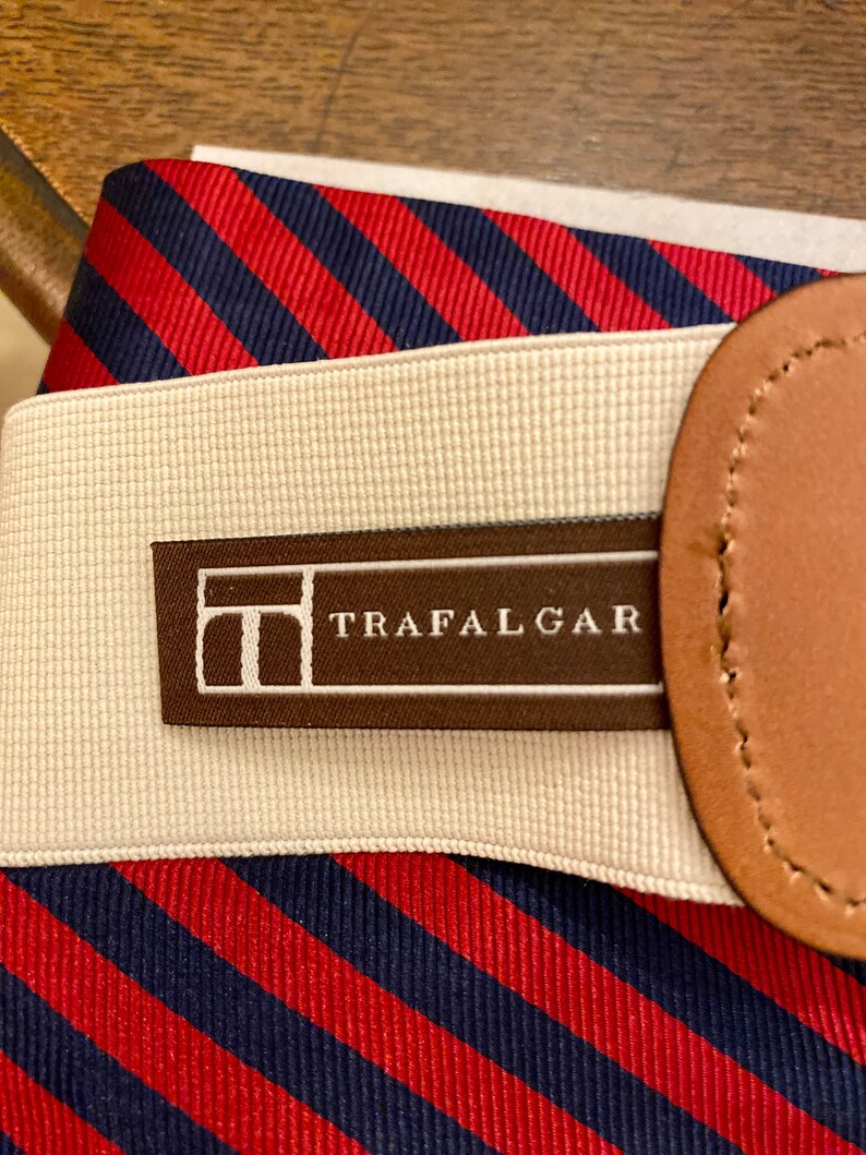 May include: Close-up of a striped belt with the word "TRAFALGAR" printed on a brown label. The belt features red and navy blue diagonal stripes, a beige elastic band, and a brown leather detail.