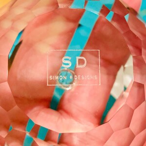 K&ouml;nnte beinhalten: Eine klare, facettierte Kristallkugel mit dem Text "SD SIMON DESIGNS" auf der Oberfl&auml;che. Die Kugel hat ein geometrisches Muster und reflektiert Licht, mit einem t&uuml;rkisfarbenen Band, das durch den Kristall sichtbar ist.