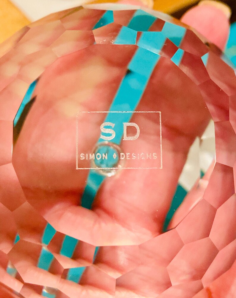 K&ouml;nnte beinhalten: Eine klare, facettierte Kristallkugel mit dem Text "SD SIMON DESIGNS" auf der Oberfl&auml;che. Die Kugel hat ein geometrisches Muster und reflektiert Licht, mit einem t&uuml;rkisfarbenen Band, das durch den Kristall sichtbar ist.