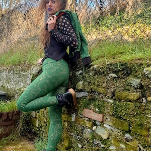 Puede incluir: Una mujer con leggings verdes con un patrón dorado, un chaleco negro y una chaqueta verde. Está de pie junto a un muro de piedra con musgo creciendo en él.