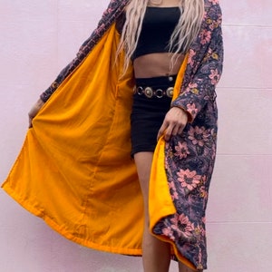 K&ouml;nnte beinhalten: Eine lange Robe mit Blumenmuster und gelbem Futter. Die Robe ist offen und die Person, die sie tr&auml;gt, steht vor einer rosa Wand. Die Person tr&auml;gt ein schwarzes Crop-Top und schwarze Shorts.