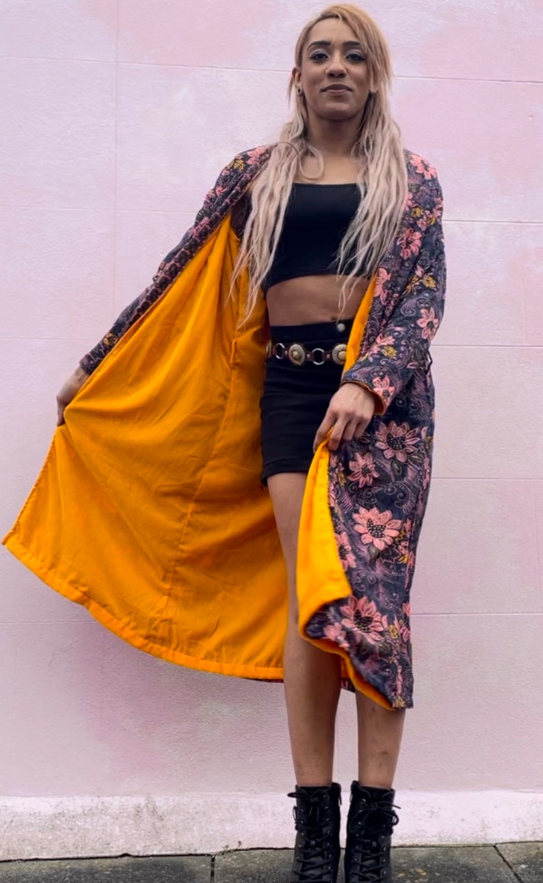 K&ouml;nnte beinhalten: Eine lange Robe mit Blumenmuster und gelbem Futter. Die Robe ist offen und die Person, die sie tr&auml;gt, steht vor einer rosa Wand. Die Person tr&auml;gt ein schwarzes Crop-Top und schwarze Shorts.