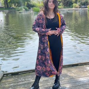 K&ouml;nnte beinhalten: Eine lange Robe mit floralem Muster und gelbem Futter. Die Robe wird &uuml;ber einem schwarzen Tanktop und einer schwarzen Hose getragen. Die Person steht auf einem Holzsteg an einem Teich.