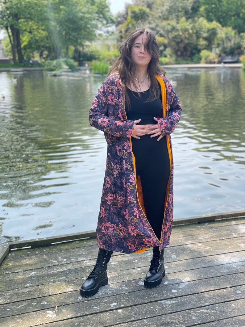 K&ouml;nnte beinhalten: Eine lange Robe mit floralem Muster und gelbem Futter. Die Robe wird &uuml;ber einem schwarzen Tanktop und einer schwarzen Hose getragen. Die Person steht auf einem Holzsteg an einem Teich.