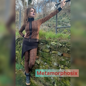 May include: A woman wearing a brown and black geometric patterned jacket and matching leggings. She is standing in front of a brick wall with green moss growing on it. The text "Metamorphosix" is visible in the lower right corner of the image.