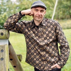 Puede incluir: Un hombre con una camisa de manga larga con estampado negro y dorado y una gorra de golf gris. Se apoya en una valla de madera.