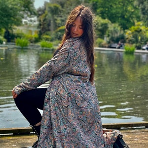 Peut inclure: Longue veste en tissu imprimé cachemire dans les tons de bleu, orange et violet. La veste est portée avec un pantalon et des bottes noirs. La scène se déroule en extérieur, près d'un plan d'eau.