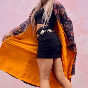 K&ouml;nnte beinhalten: Eine Person tr&auml;gt ein schwarzes Crop-Top, einen schwarzen Minirock und eine lange, floral gemusterte Robe mit gelbem Futter. Die Robe ist offen und das Futter ist sichtbar. Die Person steht vor einer rosa Wand.