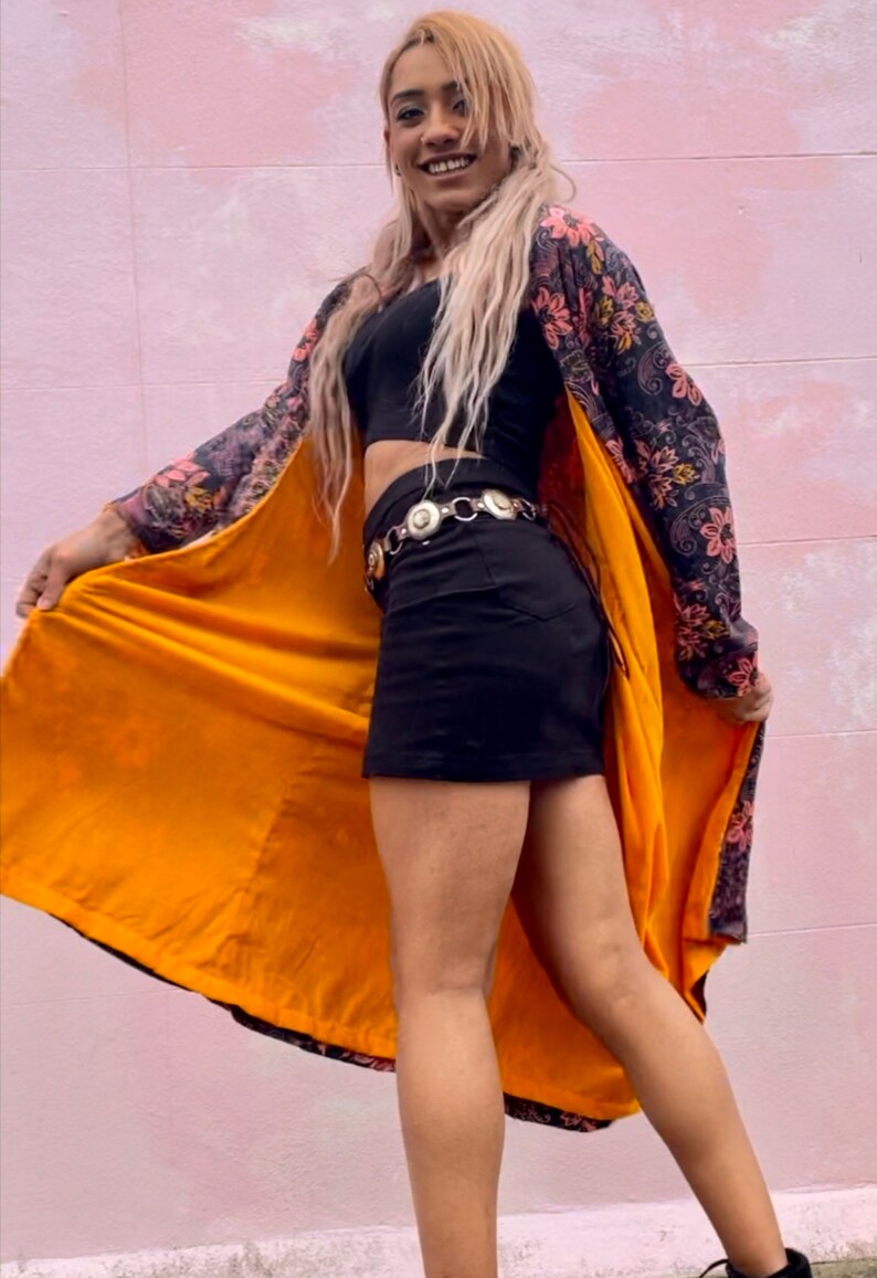 K&ouml;nnte beinhalten: Eine Person tr&auml;gt ein schwarzes Crop-Top, einen schwarzen Minirock und eine lange, floral gemusterte Robe mit gelbem Futter. Die Robe ist offen und das Futter ist sichtbar. Die Person steht vor einer rosa Wand.