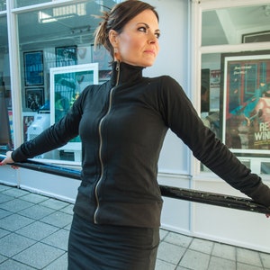 Pode incluir: Uma mulher vestindo um top preto de gola alta com z&iacute;per e uma saia preta com fenda. Ela est&aacute; em p&eacute; na frente de uma janela com cartazes nela.
