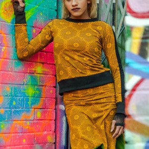 Pode incluir: Uma mulher vestindo um top e uma saia com padrão amarelo e preto com uma faixa preta na cintura. Ela está em pé na frente de uma parede de tijolos colorida com grafite.