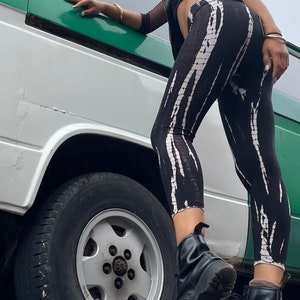 Può includere: Una donna con un top corto nero, un giubbotto di pelle nero e leggings a righe bianche e nere si trova accanto a un furgone bianco con una striscia verde. Indossa stivali neri e ha i capelli tirati indietro.
