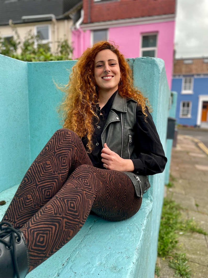 May include: A woman with long, curly red hair sits on a blue bench wearing a black leather vest and brown patterned leggings.
