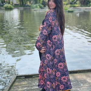 K&ouml;nnte beinhalten: Eine lange, flie&szlig;ende Robe mit Blumenmuster auf dunkelblauem Grund und rosa und gelben Bl&uuml;ten. Die Robe wird von einer Person getragen, die auf einem Holzsteg an einem Teich steht.