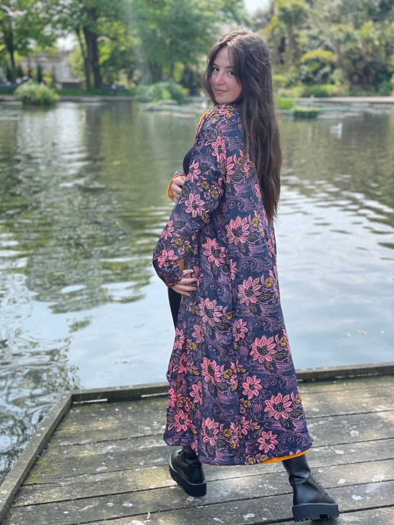 K&ouml;nnte beinhalten: Eine lange, flie&szlig;ende Robe mit Blumenmuster auf dunkelblauem Grund und rosa und gelben Bl&uuml;ten. Die Robe wird von einer Person getragen, die auf einem Holzsteg an einem Teich steht.