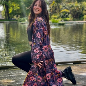K&ouml;nnte beinhalten: Eine Frau tr&auml;gt einen langen, floral gemusterten Kimono mit einem schwarzen Crop-Top und schwarzen Leggings. Sie kniet auf einem Holzsteg mit einem Teich im Hintergrund.